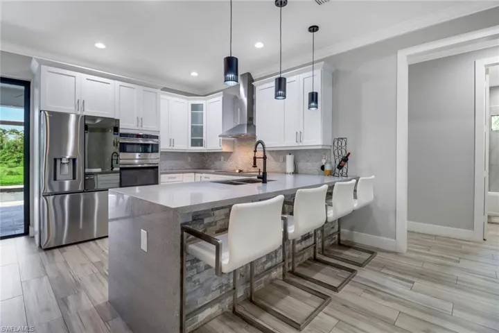Kitchen featuring stainless steel appliances, wall chimney range hood, hanging light fixtures, backsplash, and white cabinetry