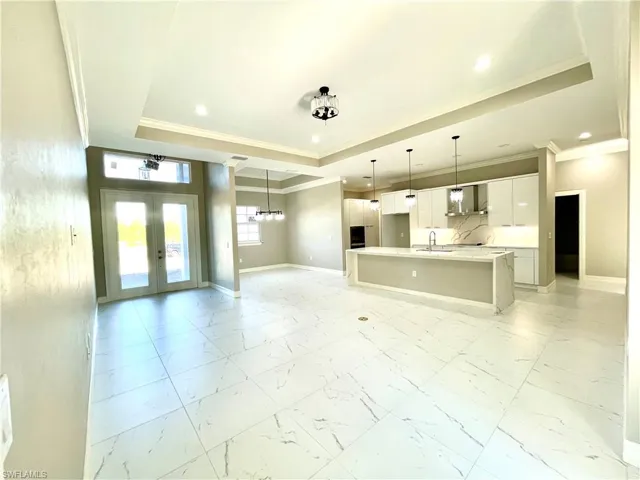 Unfurnished living room with ornamental molding, light marble finish flooring, a chandelier, a raised ceiling, and french doors