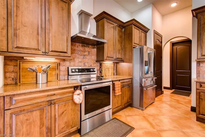 Kitchen featuring wall chimney range hood, light tile patterned floors, tasteful backsplash, light stone counters, and stainless steel appliances