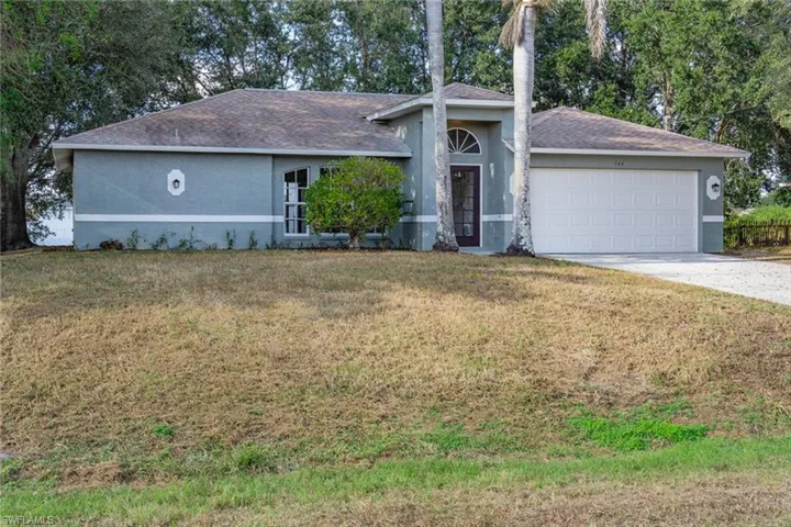 Single story home featuring stucco siding, a front lawn, roof with shingles, driveway, and view of scattered trees