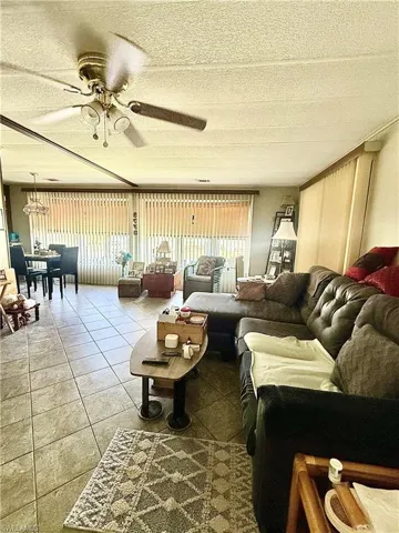 Spacious living area featuring tile flooring throughout, a brass-finish ceiling fan with lighting, ample natural light from large windows with vertical blinds, and distinct dining and seating zones