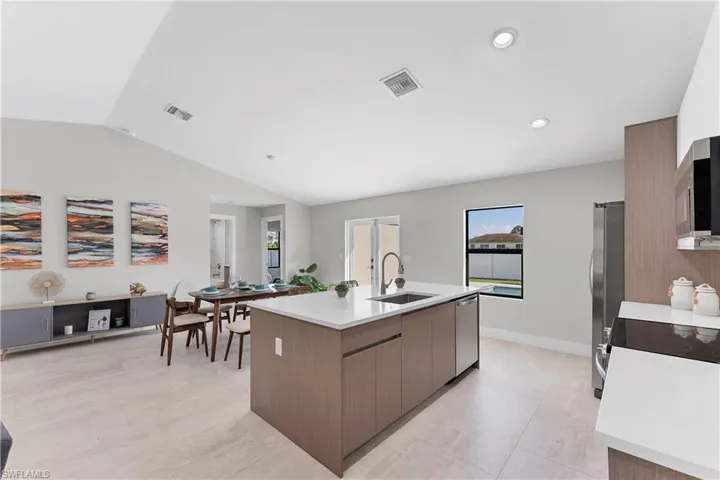 Kitchen featuring a center island with a stone-finish countertop and stainless steel sink, wood-finish cabinetry, recessed lighting, and light-toned tile flooring under a vaulted ceiling