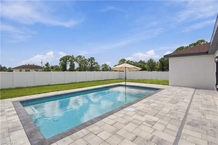 In-ground swimming pool with a dark stone-finish border, expansive paver patio, and white vinyl privacy fencing