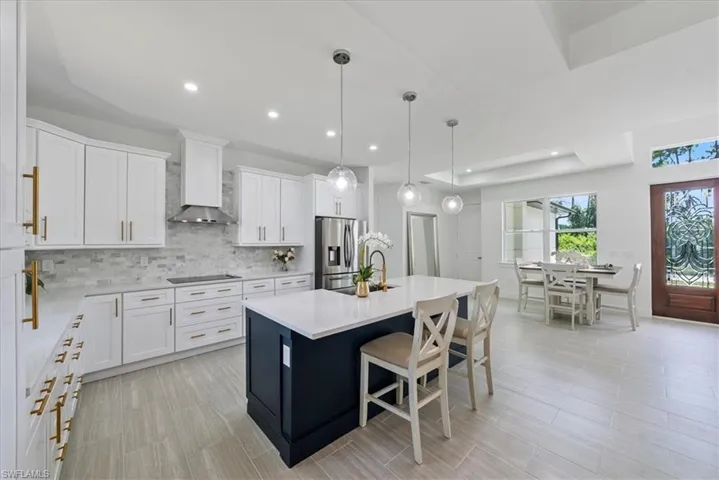 Kitchen with white cabinetry, hanging light fixtures, an island with sink, wall chimney exhaust hood, and stainless steel fridge