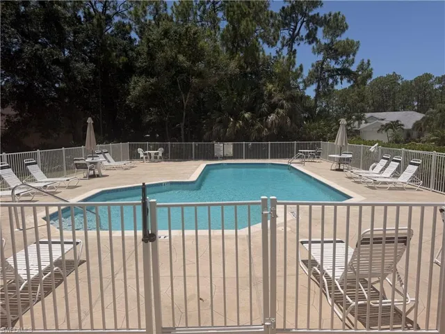 Fenced pool area featuring a freeform swimming pool, textured concrete decking, white perimeter fencing, and surrounding mature trees