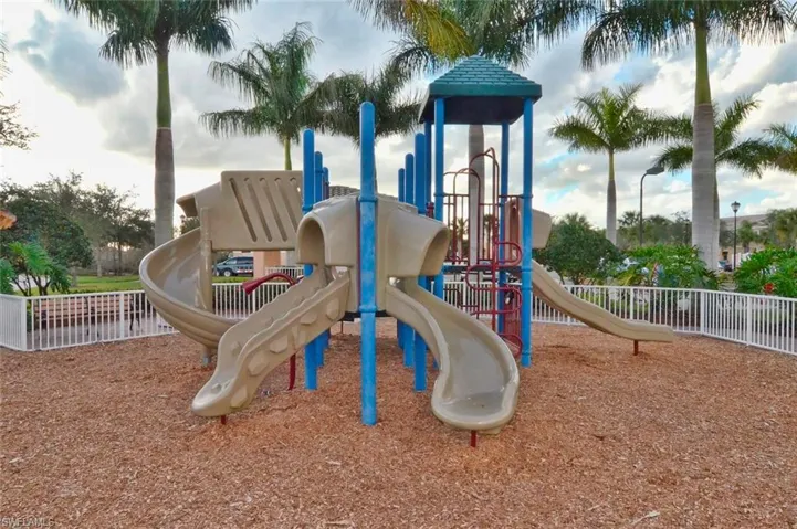 Playground structure with multiple slides and a climbing apparatus set on a wood chip base, enclosed by a white picket fence
