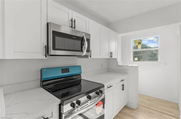 White shaker cabinetry with matte black hardware, stainless steel microwave, electric range, light-toned wood-finish flooring, and a window providing natural light