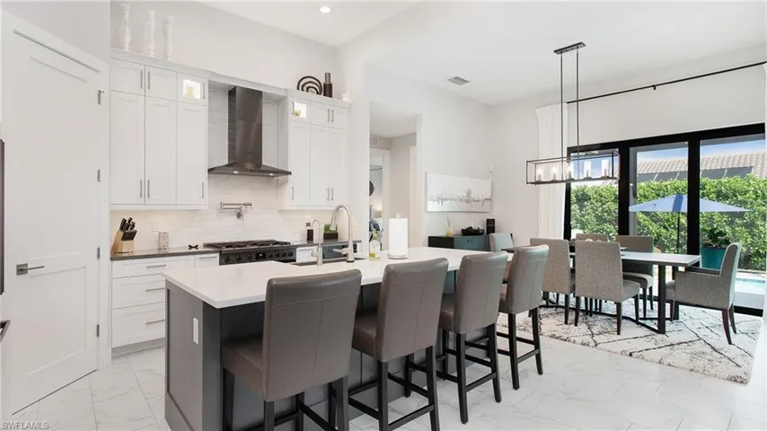 Kitchen with white cabinets, decorative backsplash, an island with sink, wall chimney range hood, and pendant lighting