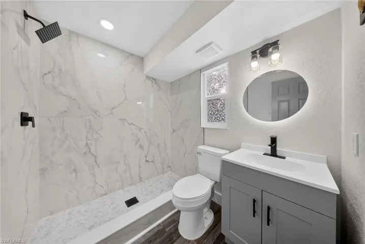 Bathroom featuring a walk-in shower with large format marble-look tile, matte black shower fixtures, a gray vanity with white countertop, and an illuminated oval mirror