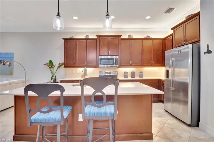 Kitchen featuring appliances with stainless steel finishes, a center island with sink, light tile patterned floors, hanging light fixtures, and brown cabinetry