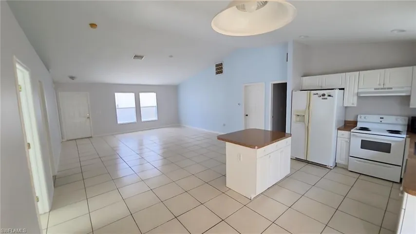 Expansive open concept interior featuring light tile flooring throughout, vaulted ceilings, two large windows, a built-in kitchen island with cabinetry, and white kitchen appliances