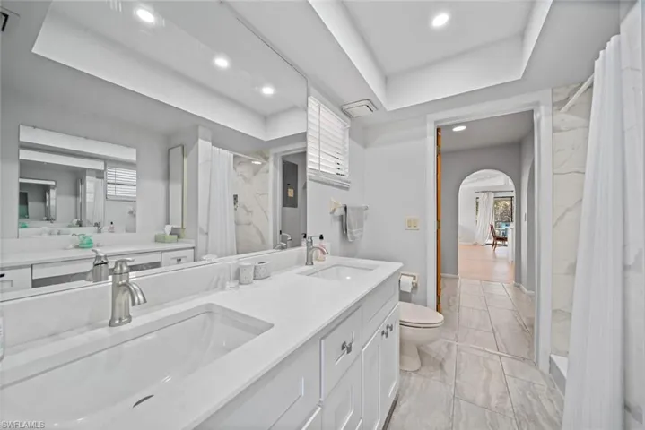 Full bathroom featuring a raised ceiling, toilet, marble look tiles, double vanity, and recessed lighting