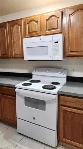 Kitchen featuring white appliances, wood finish cabinetry, dark countertops, and light tile patterned flooring
