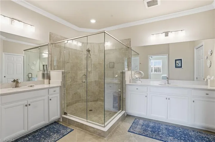 Primary bathroom with crown molding, dual vanities, spacious walk-in shower, patterned tile floors, ample space, and linen closet.