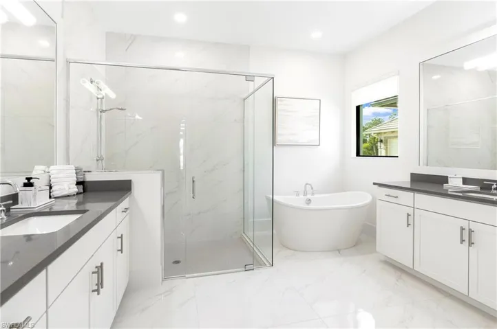 Full bath featuring two vanities, a marble finish shower, a freestanding tub, light marble finish flooring, and recessed lighting