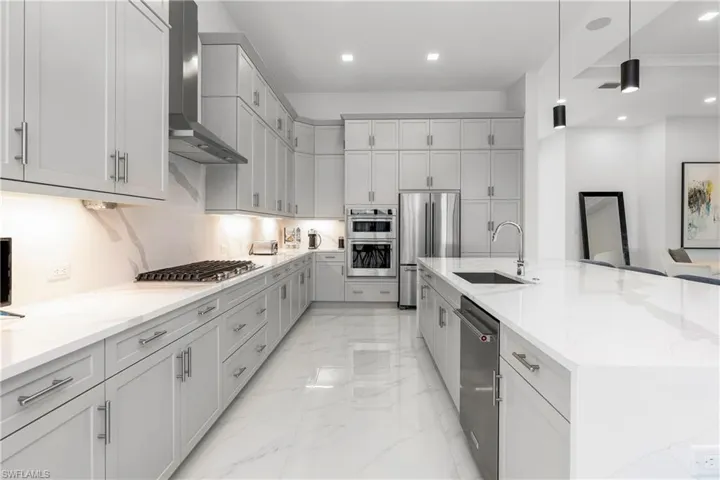 Kitchen featuring wall chimney range hood, decorative light fixtures, light stone counters, appliances with stainless steel finishes, and a large island with sink
