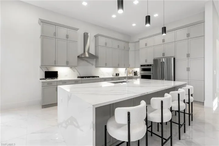Kitchen featuring gray cabinetry, light stone counters, wall chimney exhaust hood, a kitchen bar, and stainless steel appliances