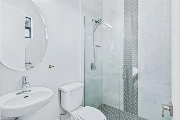 Bathroom featuring a glass-enclosed shower with a hexagon tile floor, a contemporary pedestal sink, a circular mirror, and white tile walls