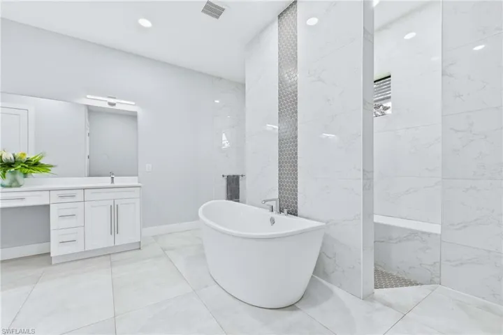 Spacious bathroom featuring a freestanding soaking tub, large format tile flooring, a walk-in shower with a built-in bench, a double vanity with a mirrored backsplash, and recessed lighting
