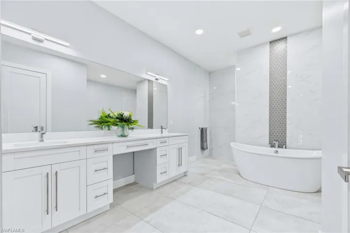 Spacious bathroom featuring a freestanding soaking tub, dual vanity with white cabinetry, large format floor tiling, and a tiled accent wall with hexagonal details