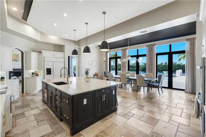 Two tone kitchen with two tone color scheme, hanging light fixtures, light stone countertops, stone tile floors, and an island with sink