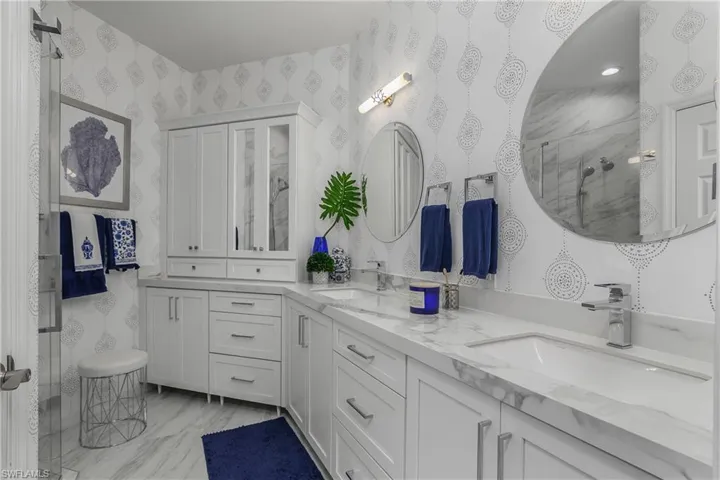 Primary Bathroom with double vanity, a marble finish shower, wallpapered walls, and light marble finish flooring