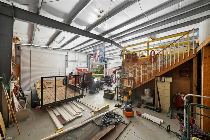 Spacious garage interior featuring industrial-style steel beam construction, two large roll-up doors, concrete flooring, a built-in wooden staircase with a banister, and a loft area with a yellow safety railing