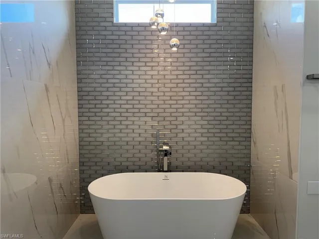 Freestanding white soaking tub with a floor-mounted polished chrome faucet, accented by a grey brick-tiled feature wall, large-format veined tile surround, and three-globe pendant lighting