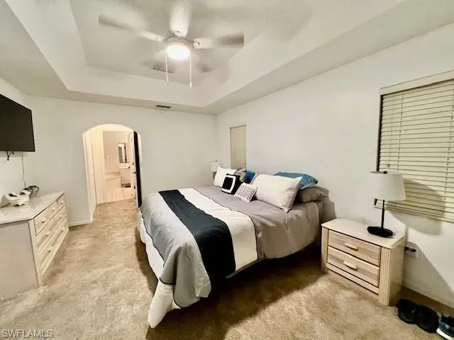 Bedroom featuring a tray ceiling, light colored carpet, arched walkways, and ceiling fan