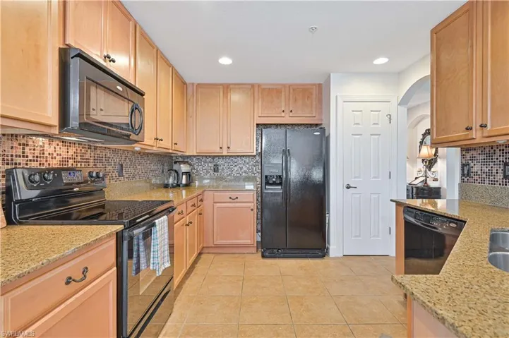 Kitchen with black appliances, light stone counters, light wood finish cabinets, light tile patterned floors, and arched walkways