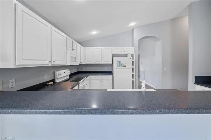 Kitchen with dark countertops, vaulted ceiling, white appliances, white cabinets, and recessed lighting