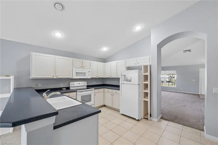 Kitchen featuring dark countertops, white appliances, white cabinetry, light colored carpet, and vaulted ceiling
