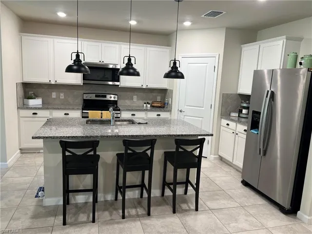 Kitchen featuring light stone counters, hanging light fixtures, a center island with sink, appliances with stainless steel finishes, and white cabinets