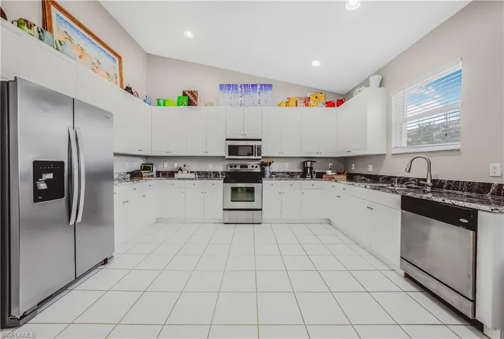 Kitchen featuring stainless steel appliances, dark stone counters, vaulted ceiling, white cabinets, and light tile patterned floors