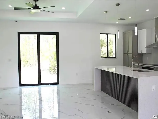 Spacious open concept interior featuring large format marble-look tile flooring, a modern kitchen island with textured paneling and white countertop, recessed lighting, and a ceiling fan