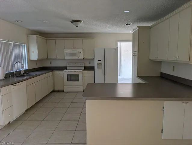 Kitchen with white appliances, a peninsula, a textured ceiling, light tile patterned flooring, and dark countertops