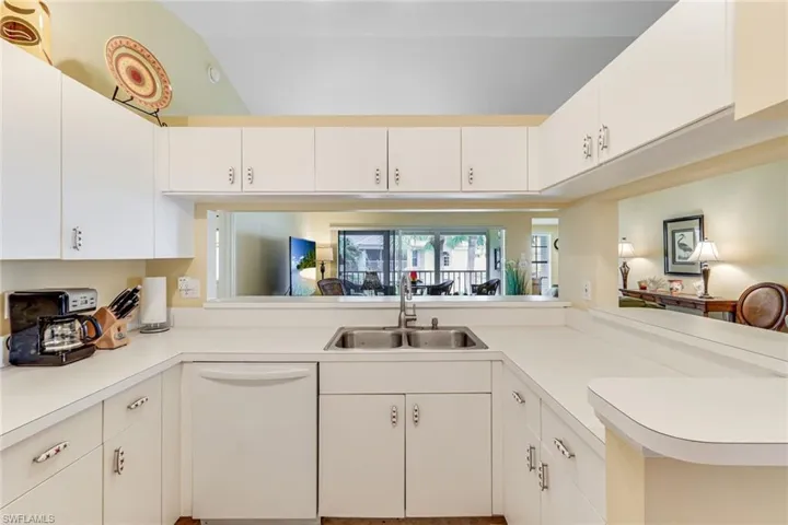 U-shaped kitchen with white cabinetry and countertops, featuring a double basin stainless steel sink and a pass-through window to the living area