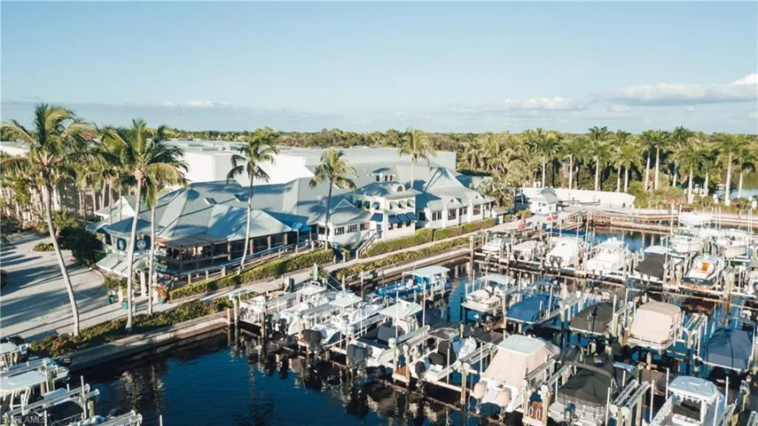 Marina featuring numerous boat slips, a multi-level building with gray roofing, surrounding palm trees, and clear blue water