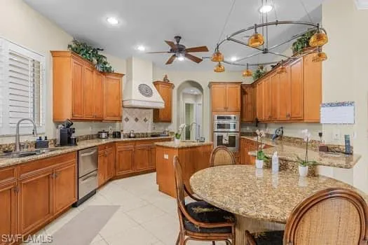 Kitchen with a kitchen island, sink, custom exhaust hood, appliances with stainless steel finishes, and ceiling fan with notable chandelier