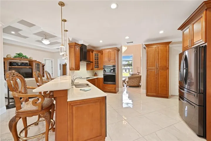 Kitchen featuring wall chimney range hood, coffered ceiling, kitchen peninsula, stainless steel appliances, and hanging light fixtures