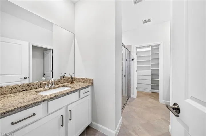 Bathroom featuring white cabinetry, stone-surface countertop with an undermount sink, full-width mirror, glass-enclosed shower, and built-in walk-in closet shelving