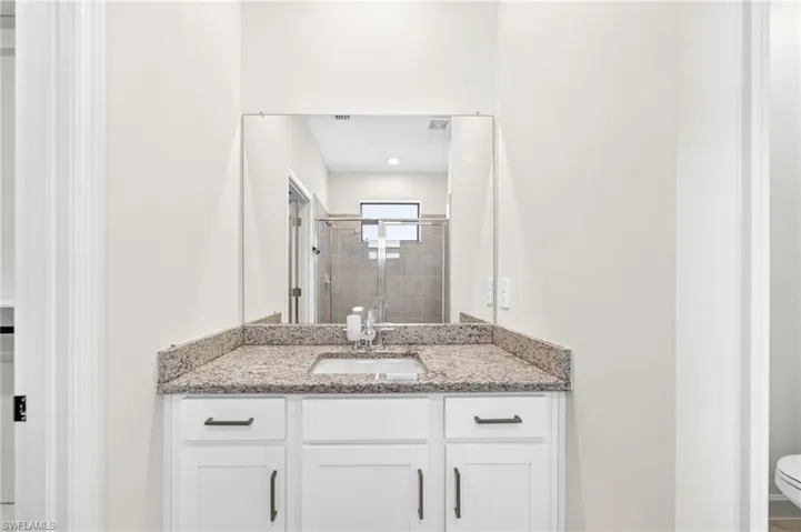 White cabinetry with dark hardware, stone-composite countertop, undermount sink, expansive wall mirror, and tiled shower enclosure with glass door