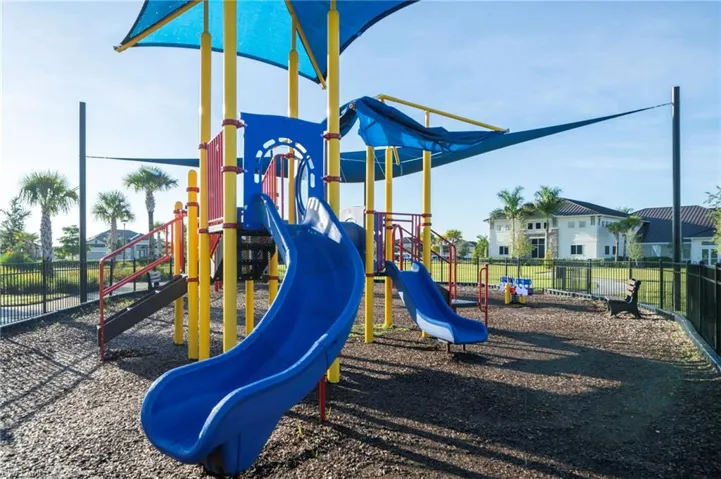 Outdoor recreational area featuring a multi-level climbing structure with integrated slides, shade canopy, fenced perimeter, and wood chip surfacing