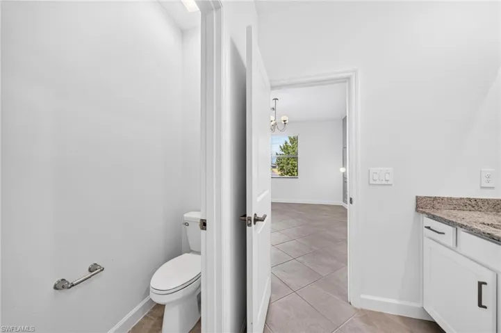 Neutral-toned tile flooring, white shaker-style cabinetry with stone-surface countertop, and elongated toilet with chrome hardware