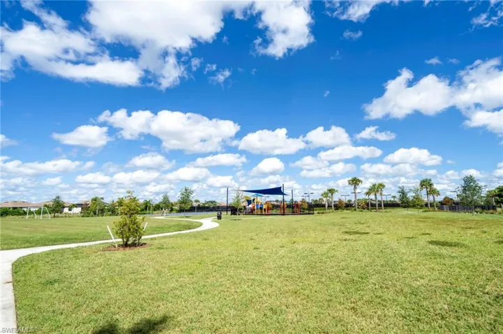 Expansive communal green space featuring a paved walkway, recreational play area with shaded structure, and palm trees