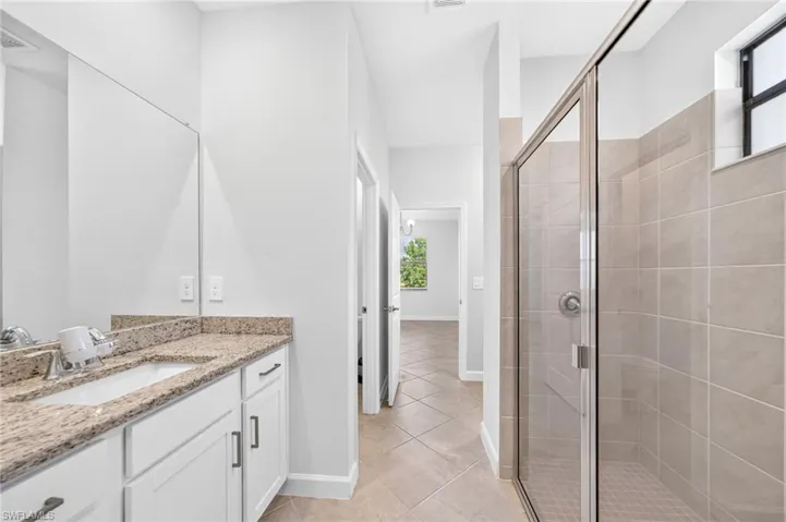 Bathroom featuring a stone-top vanity with an integrated sink, white shaker-style cabinetry, a glass-enclosed shower with tiled surround, and ceramic tile flooring