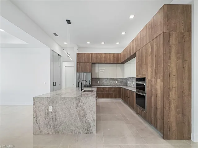 Kitchen with modern cabinets, light stone counters, backsplash, light tile patterned floors, and an island with sink