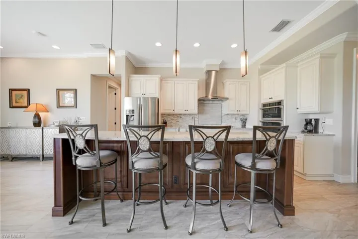 Kitchen with a large island with sink, wall chimney exhaust hood, hanging light fixtures, appliances with stainless steel finishes, and light stone counters