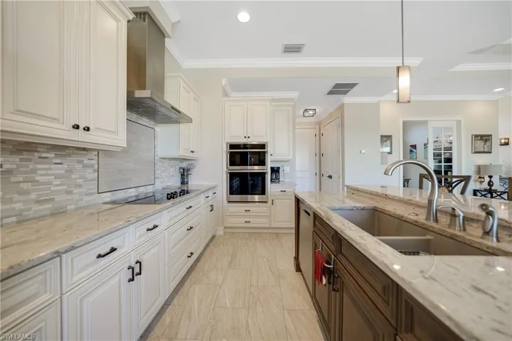 Kitchen featuring wall chimney range hood, light stone countertops, stainless steel appliances, sink, and decorative light fixtures