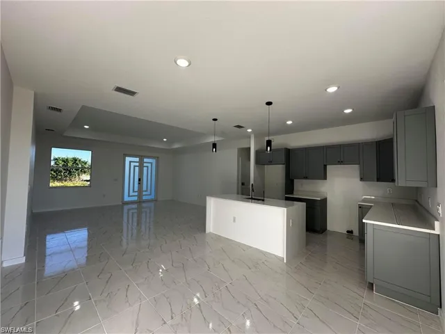 Kitchen featuring sink, gray cabinetry, decorative light fixtures, a raised ceiling, and an island with sink
